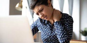 Woman sitting at a desk looking at her computer and rubbing her neck from neck pain.