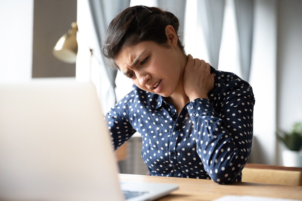 Woman sitting at a desk looking at her computer and rubbing her neck from neck pain.