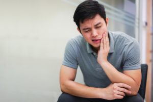 An Asian man sitting on a chair, grimacing and holding his jaw from TMJ pain.