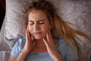 Close up view of a woman lying in bed, holding her jaw and grimacing from TMJ disorder pain.