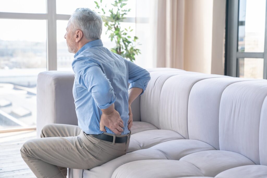 A senior man sitting on a sofa holding is lower back in pain.