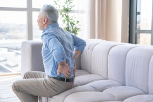 A senior man sitting on a sofa holding is lower back in pain.