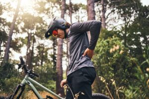 A male mountain biker stopped on his bike in the woods holding his lower back from back pain.