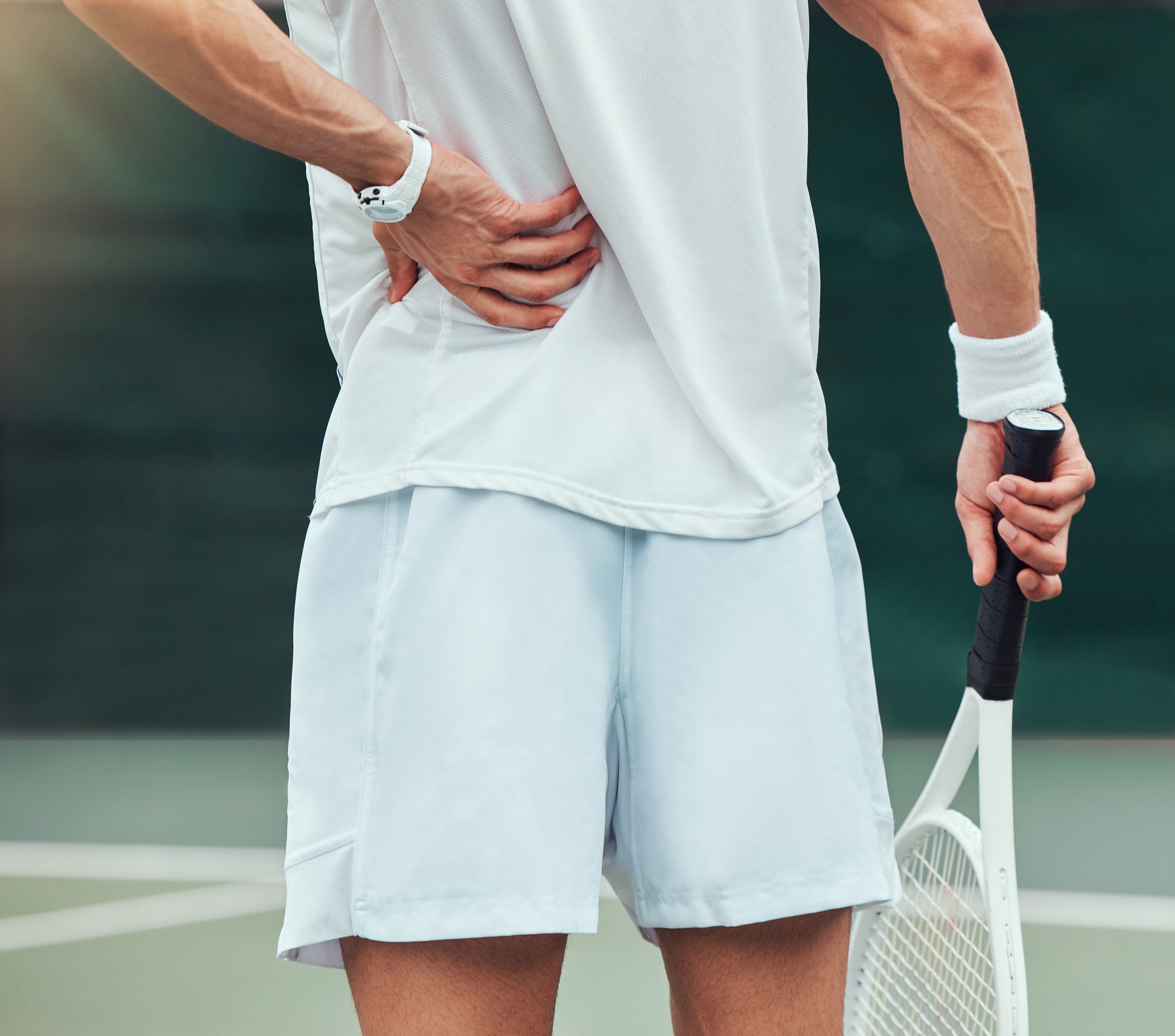 Close up of a male tennis player's backside as he holds his racquet in one hand and his lower back from back pain with the other.