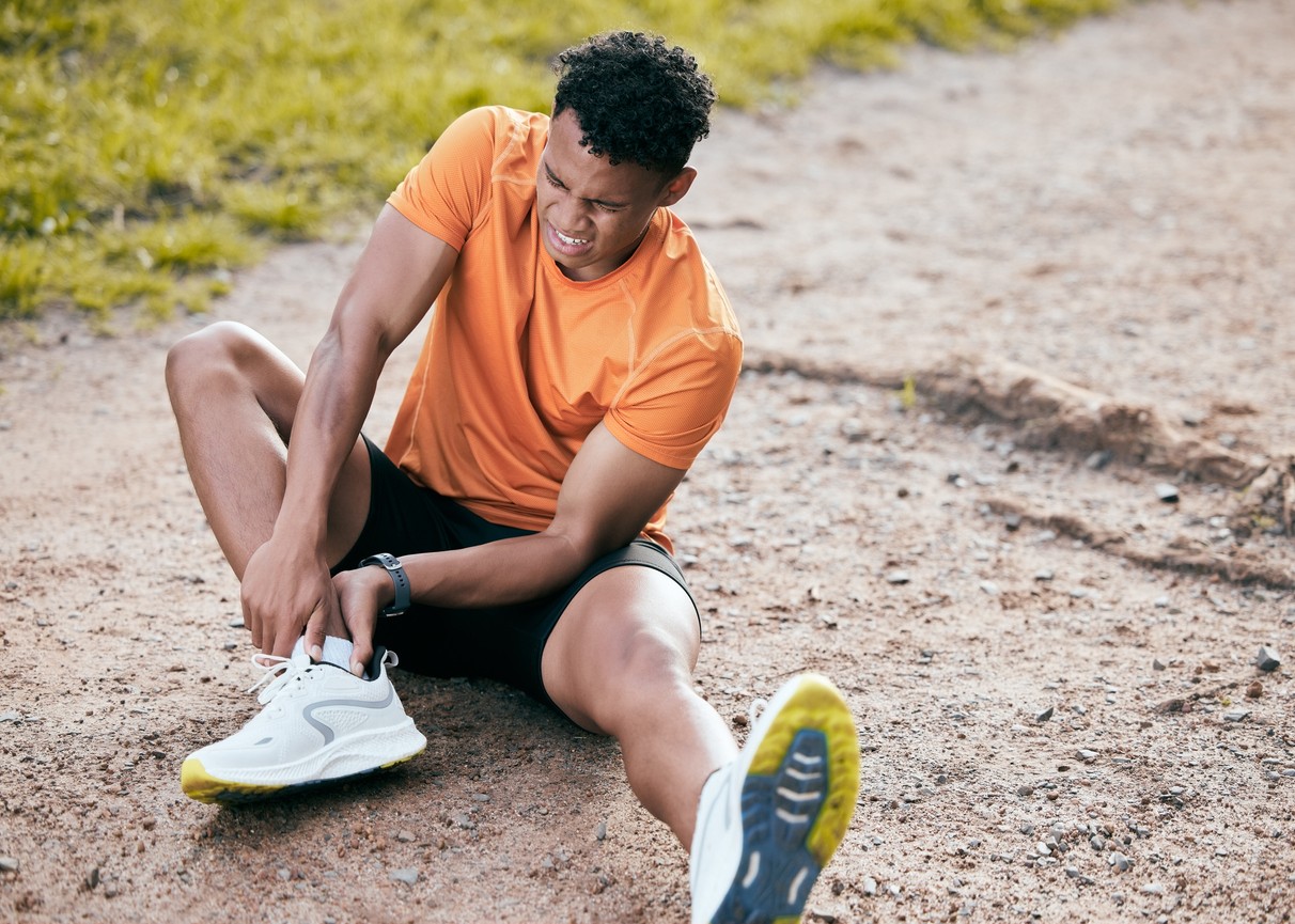African American male runner sits on gravel path holding his Achilles tendon in pain from a running injury.