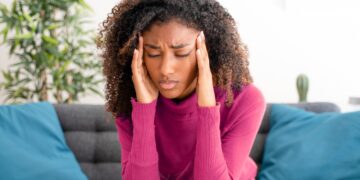 A black woman sits on the sofa and holds her head from tension headache pain.