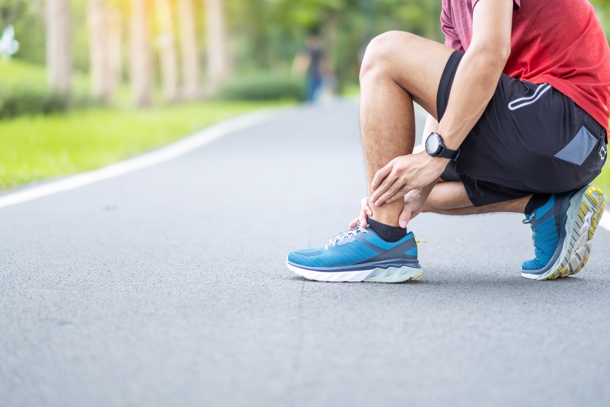 A male runner kneels on a running path to hold his achilles from tendonitis pain. 