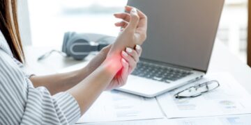 A woman sitting in front of a laptop holding her wrist in pain from tendonitis.