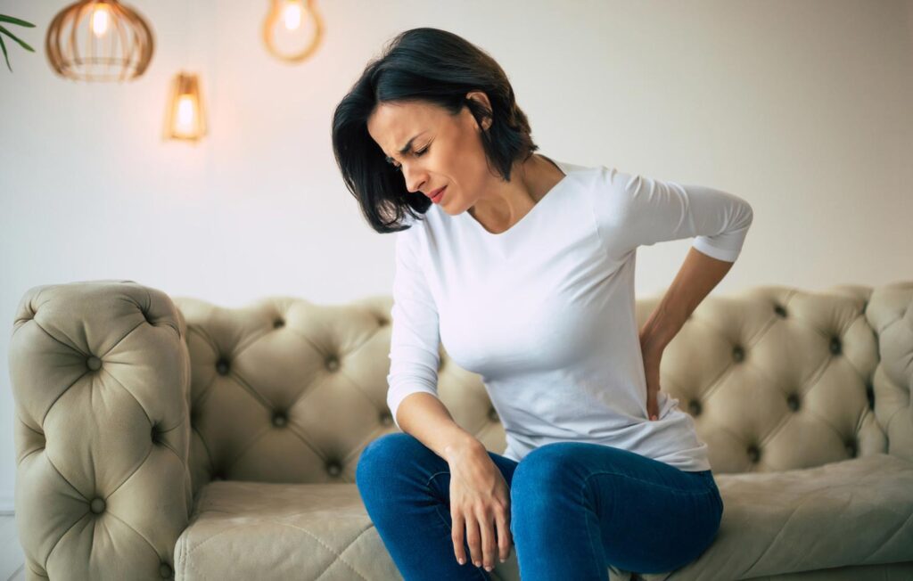 A woman with lower back pain grimaces while sitting on a sofa holding her lower back.