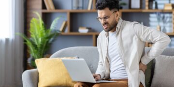 A man sits on a sofa with a laptop and holds his lower back, grimacing in pain.