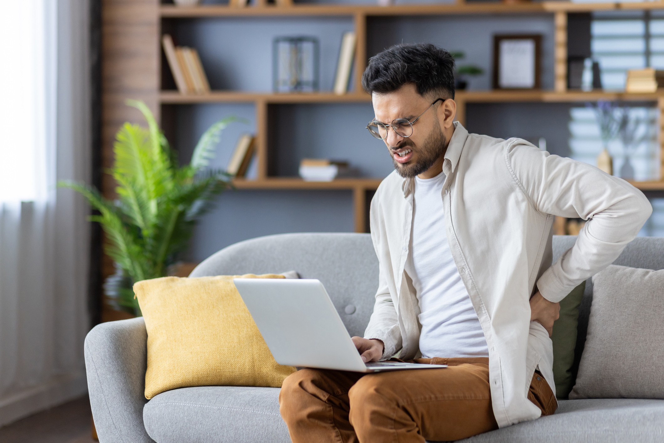 A man sits on a sofa with a laptop and holds his lower back, grimacing in pain.