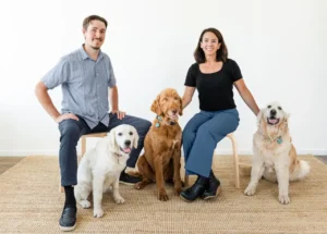 A man and a woman sit on chairs with three golden retrievers on a woven rug in a bright, minimalist room.