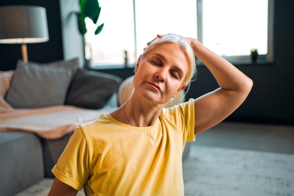 A senior woman sits and gently performs neck stretches. 