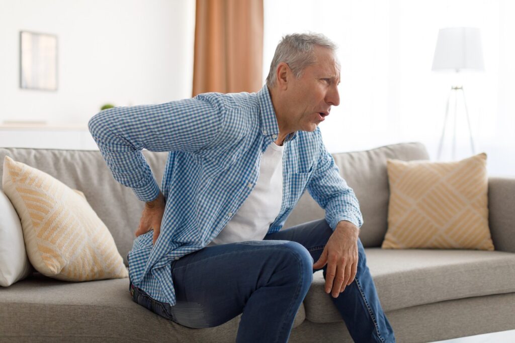 A senior man sits on the couch holding his lower back in pain from sciatica. 