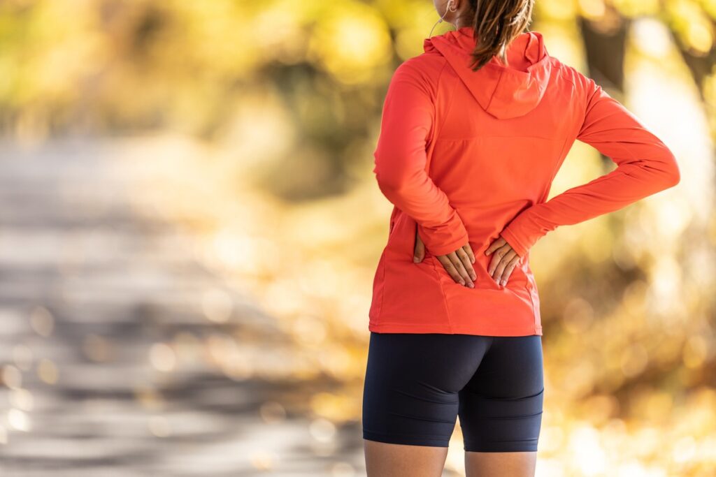 A female runner stands on a trail holding her lower back from sciatica pain. 