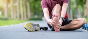 A male runner sits on the pavement holding his bare foot from plantar fasciitis pain.