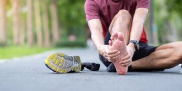 A male runner sits on the pavement holding his bare foot from plantar fasciitis pain.