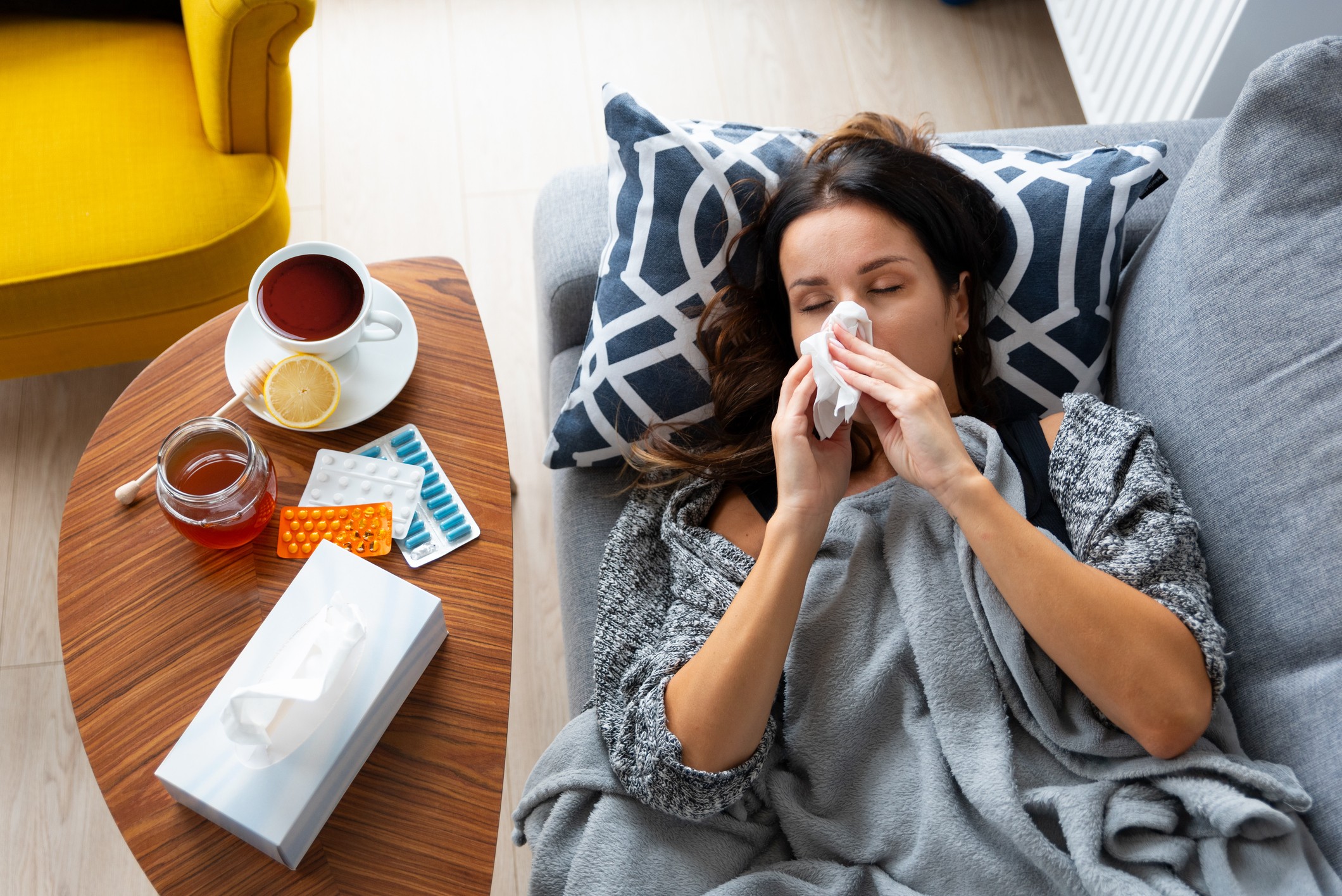 A woman lays on the couch, blowing her nose from being sick with cold and flu.