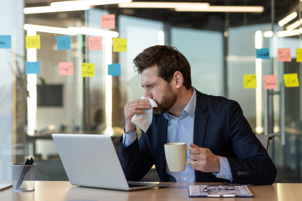 A man sneezes with a tissue in the office while holding a cup of tea at his computer.