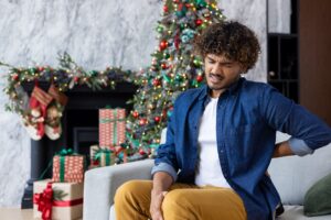 A man sits on a sofa in front of a Christmas tree grimacing with lower back pain from holiday stress.