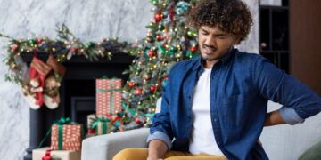 A man sits on a sofa in front of a Christmas tree grimacing with lower back pain from holiday stress.