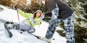 A woman sits in the snow with ski gear, while a man helps her up, both enjoying a winter day outdoors.